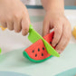 Child's hands playing with a toy watermelon and green knife on a light surface.