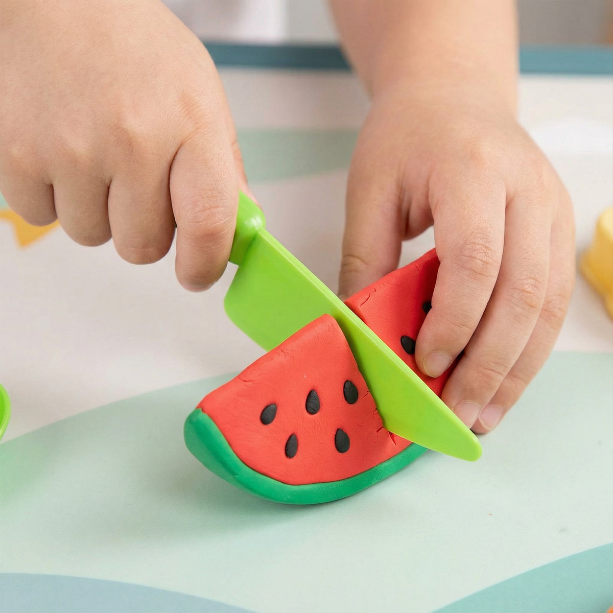 Child's hands playing with a toy watermelon and green knife on a light surface.