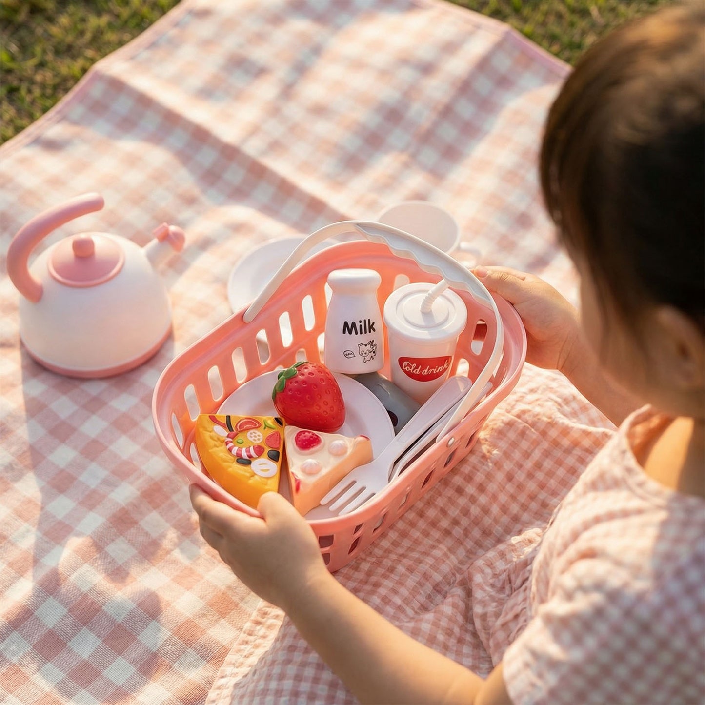 Child holding a pink toy kitchen set on a checkered blanket outdoors